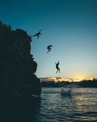 Silhouette men jumping from cliff into sea during sunset