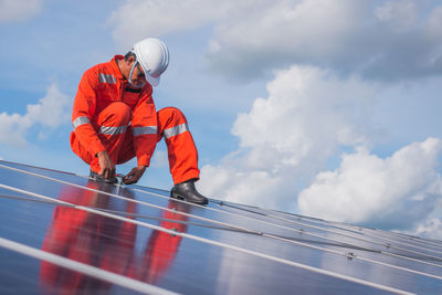 Low angle view of male technician repairing solar panels against cloudy sky