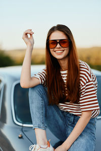 Portrait of young woman sitting on car against sky