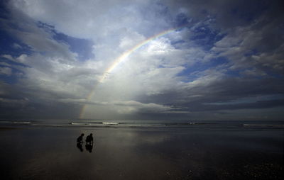 Silhouette friends at beach against rainbow in sky
