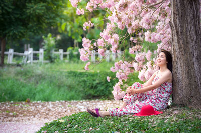 Side view of smiling woman sitting against tree trunk at public park
