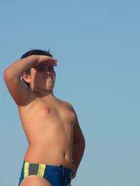 Shirtless boy shielding eyes while looking away against clear sky