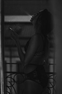 Side view of woman in lingerie smoking while standing by window at home