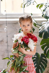 Portrait of girl holding flowers while standing against window