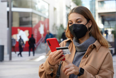 Beautiful young woman with black protective mask kn95 enters her credit card number on smart phone