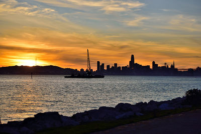 Silhouette city by sea against sky during sunset