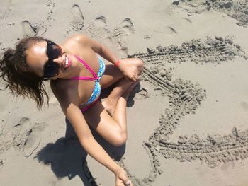 High angle portrait of woman sitting by letter m on sand at beach