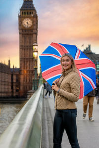 Portrait of woman standing with british flag umbrella against big ben and sky in city