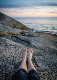 Low section of person sitting on rock by sea against sky during sunset