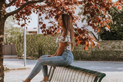 Side view of young woman sitting on tree during autumn