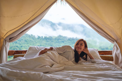 A young woman lying down on a white bed in the morning with a beautiful nature view outside the tent