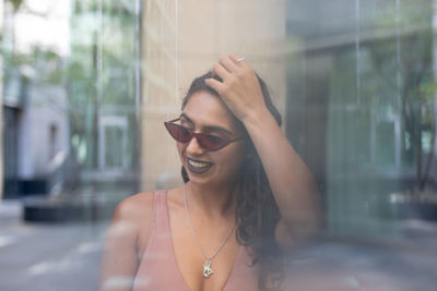 Cheerful young woman wearing sunglasses seen through glass window in city