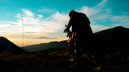 Man with motorcycle standing on mountain against sky during sunset