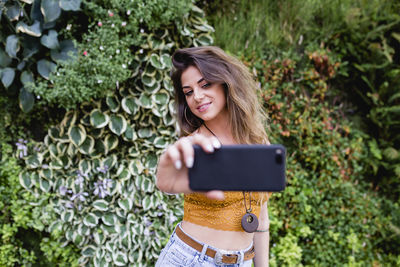 Beautiful woman taking selfie from mobile phone while standing against plants at park