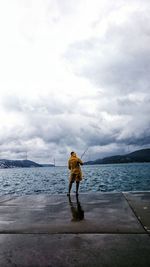 Rear view of man on pier fishing in sea against cloudy sky