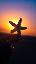 Close-up of hand holding cross against sky during sunset