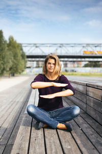 Portrait of young woman gesturing equal sign while sitting on wood against sky