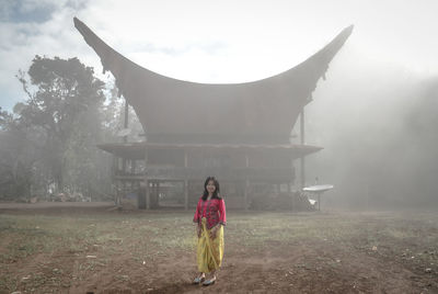 Portrait of young woman standing by tree against sky