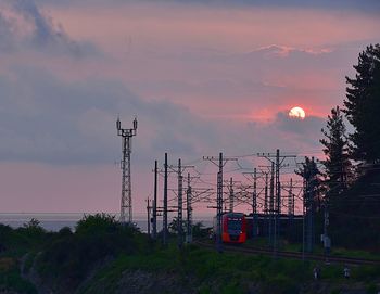 Electricity pylons by trees against sky during sunset