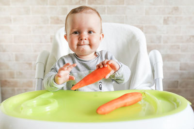 Portrait of cute baby girl with carrots sitting on high chair at home