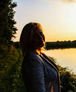 Portrait of young woman standing by lake against sky during sunset