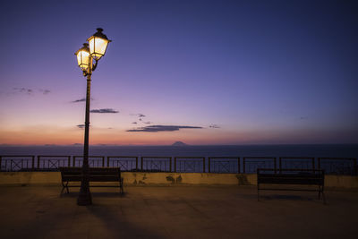 Illuminated street light by sea against sky during sunset