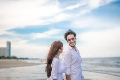 Young couple standing at beach against cloudy sky