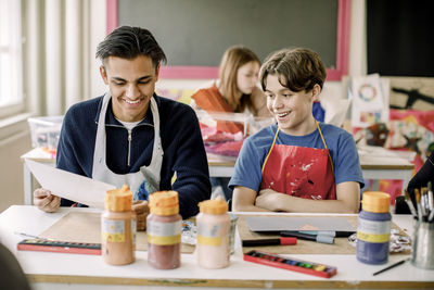 Smiling male student showing painting to teenage friend during art class at high school