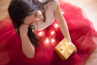 High angle view of young woman with gift and illuminated string light sitting on bed