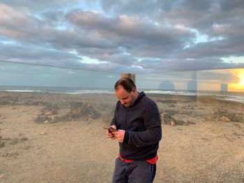 Man using mobile phone while standing on sand at beach against sky