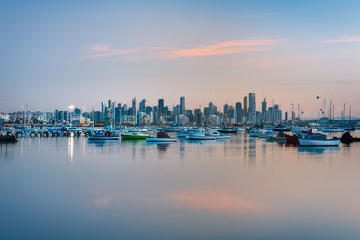 Sailboats in sea by buildings against sky during sunset