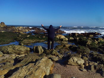 Rear view of man standing on rock at beach against blue sky