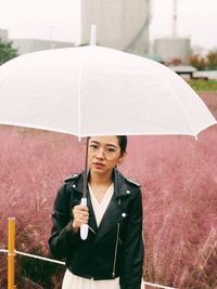 Portrait of young woman holding umbrella while standing against field in rain