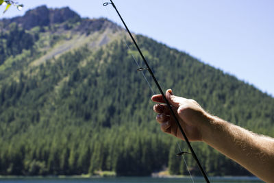 Cropped hand holding fishing rod by lake and mountains