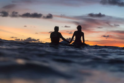 Silhouette friends in sea against cloudy sky during sunset