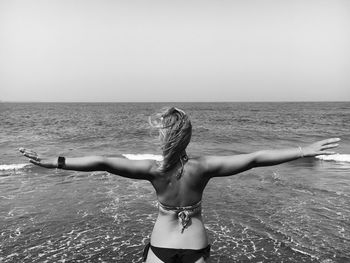 Rear view of woman in bikini standing with arms outstretched on beach against clear sky