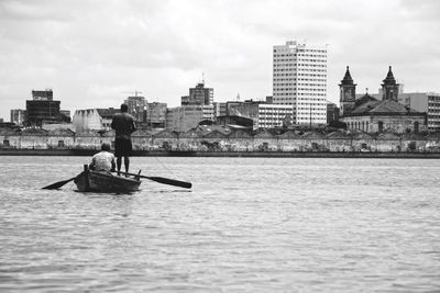 Man with friend rowing boat on river against city