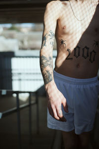 Midsection of shirtless man with tattoo in sunlight at home