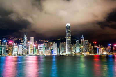 Illuminated city at waterfront against cloudy sky