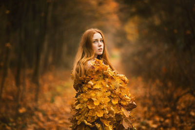 Thoughtful young woman covered with leaves standing at forest during autumn