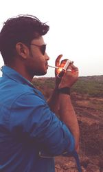 Side view of man lighting cigarette while standing against sky