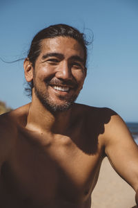 Portrait of shirtless man sitting at beach against clear blue sky during sunny day