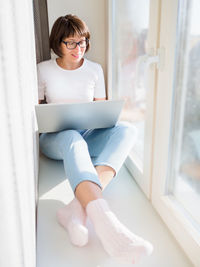 Woman works remotely from home. she sits on window sill with laptop on knees. 