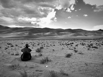 Rear view of woman sitting at desert against cloudy sky