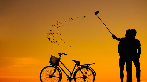 Silhouette of couple taking selfie with bicycle against sky during sunset