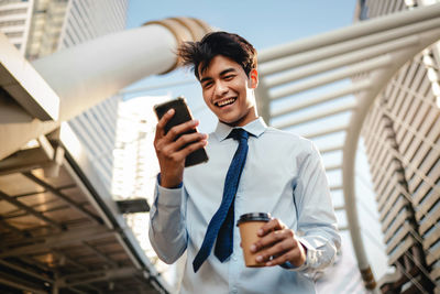 Low angle view of man wearing tie using smart phone while holding coffee outdoors