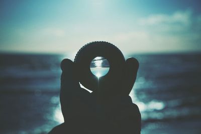 Close-up of cropped silhouette hand holding lens by sea