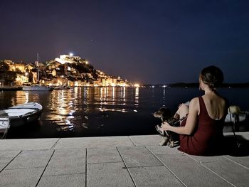 Woman with dog looking at view while sitting on promenade by sea against sky during night