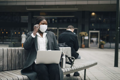 Businesswoman with laptop looking away while sitting on bench during pandemic