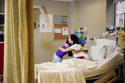 Mother embracing girl while sitting on bed in hospital